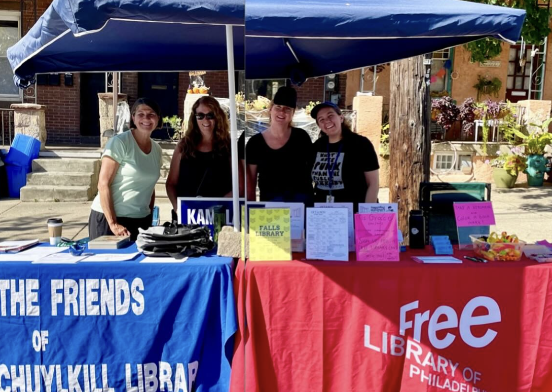 Photo take at East Falls Fall Fest showing four white women smiling behind tables with banners that read "Join the Friends of Falls of Schuylkill Library" and "Free Library of Philadelphia"